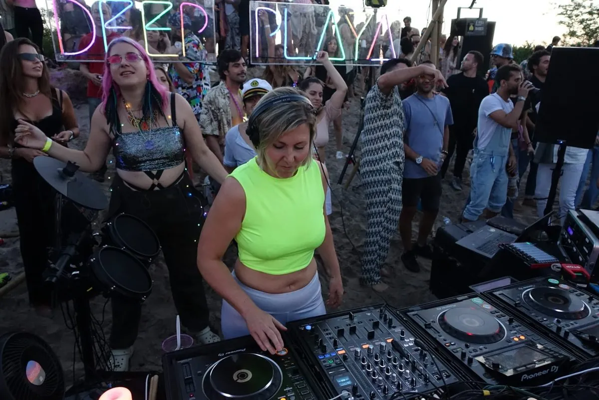 Daniela DJing outdoors beneath a Deep Playa neon sign at sunrise, surrounded by dancers