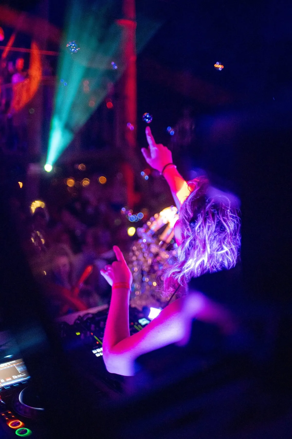 Daniela DJing with one hand raised to the crowd under purple light, with bubbles and a green laser overhead