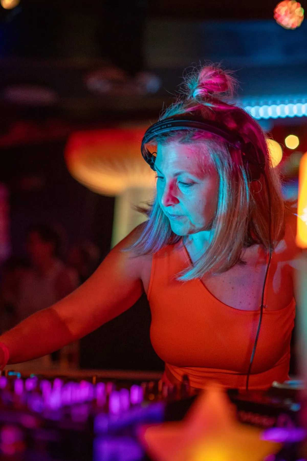 Daniela DJing in headphones and an orange top, lit in teal and magenta, with her hand on the mixer